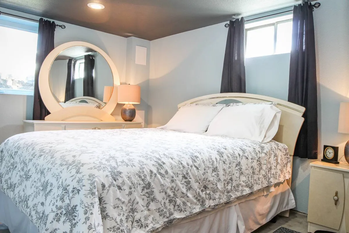 A sunlit bedroom featuring floral bedding, a round mirror above a wooden nightstand, and a cozy lamp. The room feels serene and inviting, suggesting a peaceful place to rest.