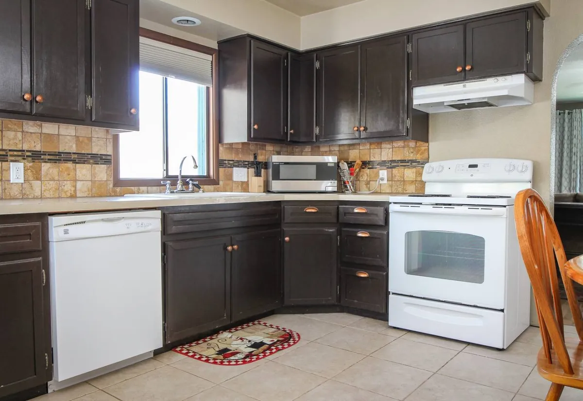 A bright, welcoming kitchen with dark wood cabinets, white appliances, a tiled backsplash, and a window letting in natural light. The space feels ready for cooking, baking, or enjoying a cup of coffee, with a layout ideal for family meals or entertaining friends.