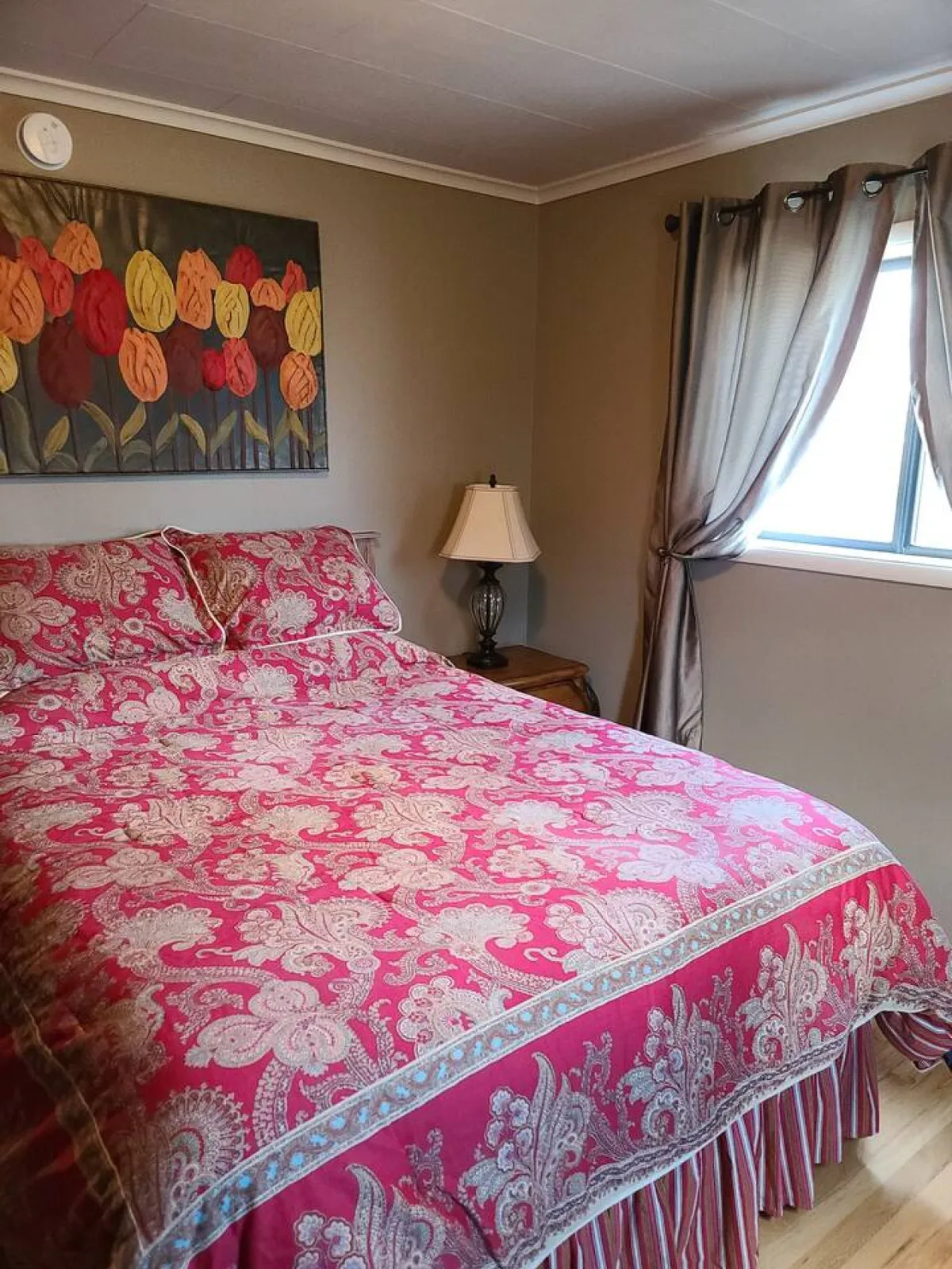 A serene queen bedroom featuring a bed with patterned bedding, colorful wall art, a nightstand with a lamp, and flowing curtains framing a window. The room exudes a peaceful and inviting mood, perfect for unwinding after a long day.