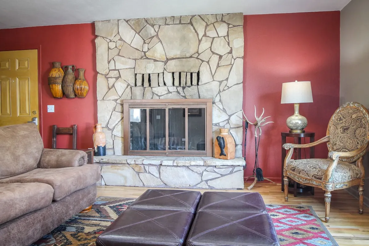 This image features a warm and welcoming living room with a stone fireplace as the focal point, a comfortable sofa, an ornate armchair, and decorative vases. The red accent wall and rich textures evoke the historic charm and inviting atmosphere described in the section.
