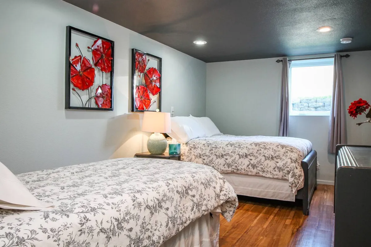 A bright, comfortable bedroom featuring two beds with floral artwork above, a lamp between them, and a window letting in gentle natural light. The space is calm and well-appointed, ideal for relaxing, reading, or sharing with a companion.