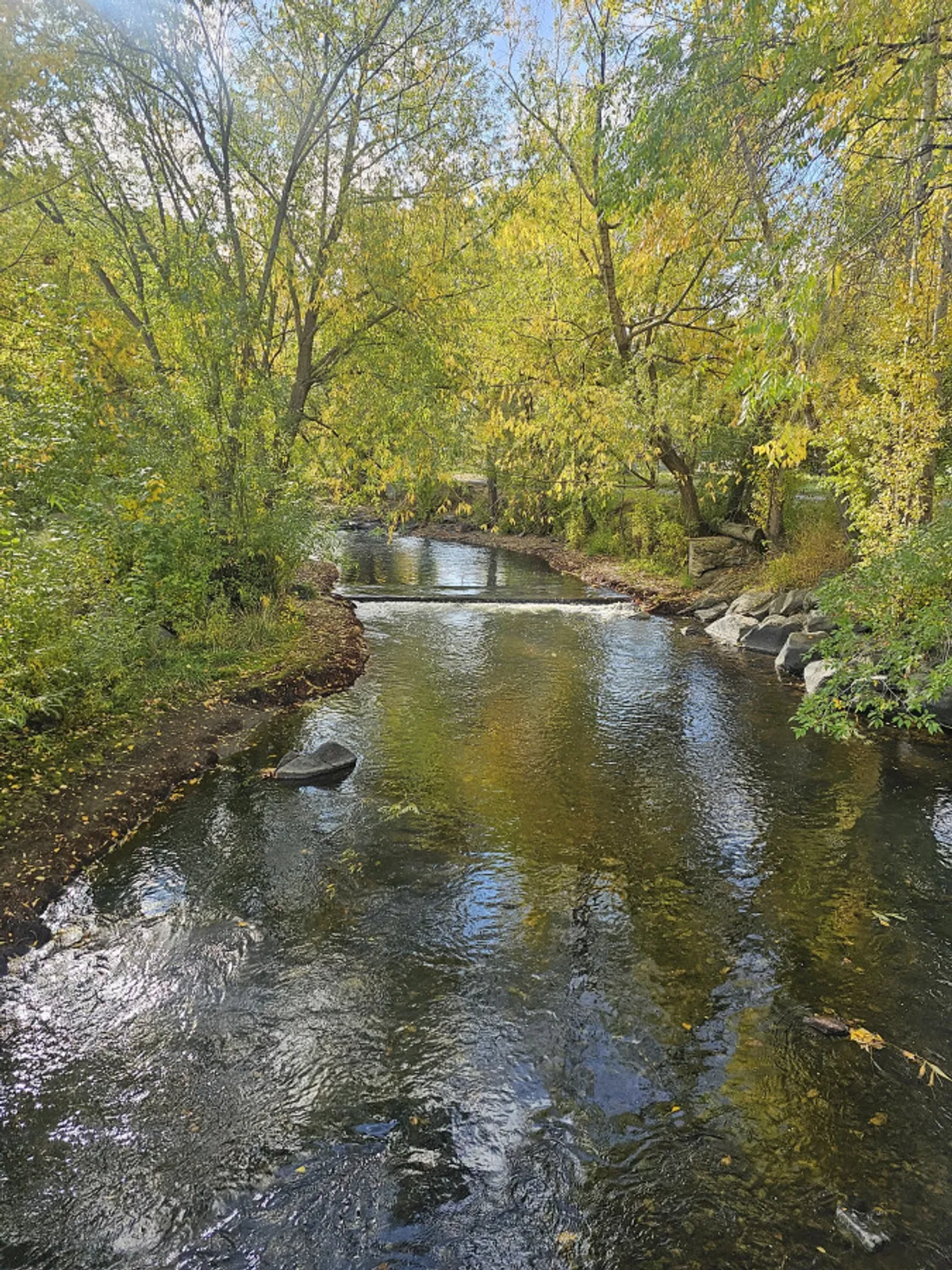 A tranquil view of a flowing river bordered by trees, with reflective water and scattered rocks creating a peaceful atmosphere. The natural setting evokes the broader outdoor attractions available in the region, such as Nordic skiing or relaxing after golf.