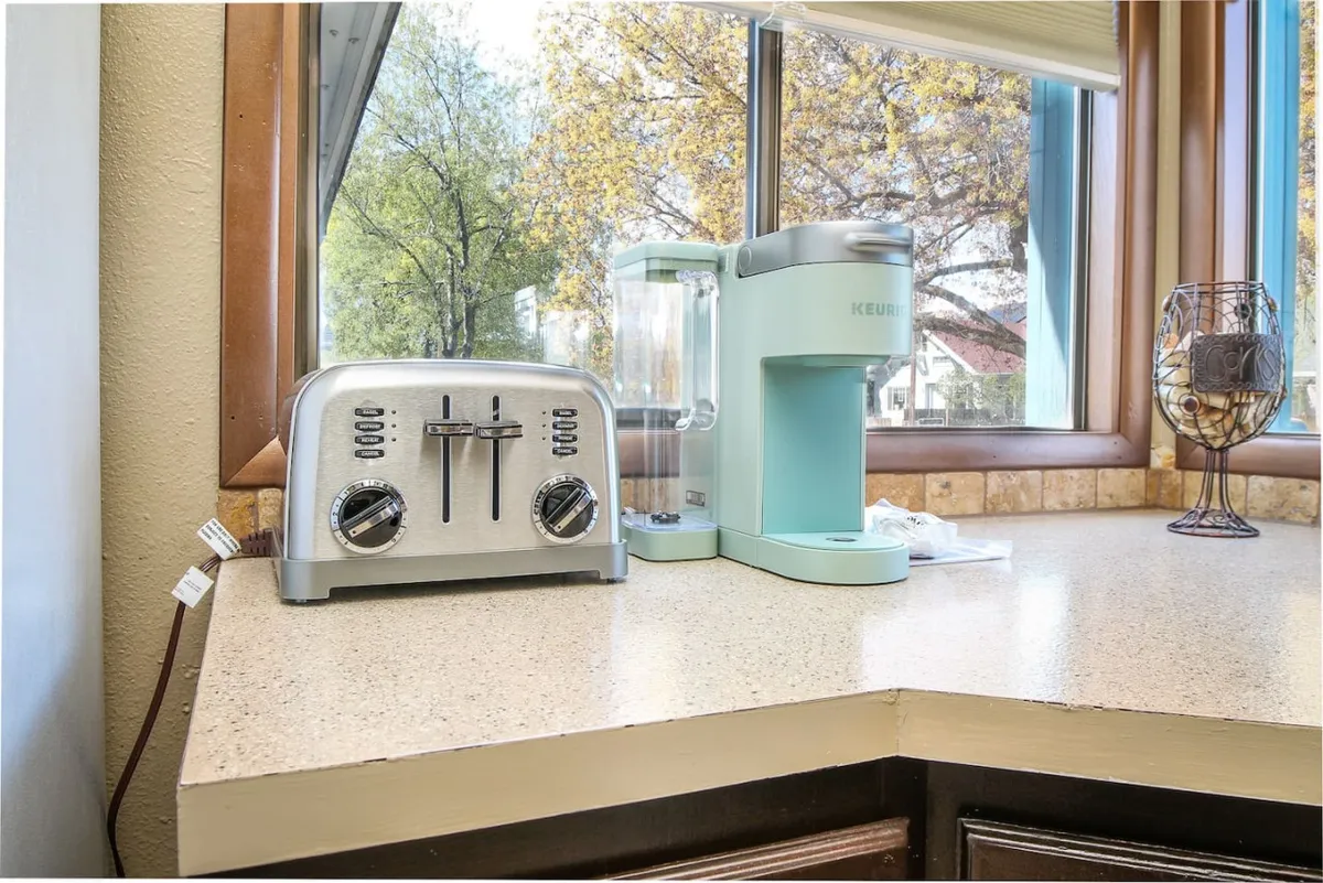 A cozy breakfast corner featuring a silver toaster, a light blue coffee maker, and large windows streaming with natural light. The space feels peaceful and inviting, ideal for enjoying coffee and simple breakfasts with a view.