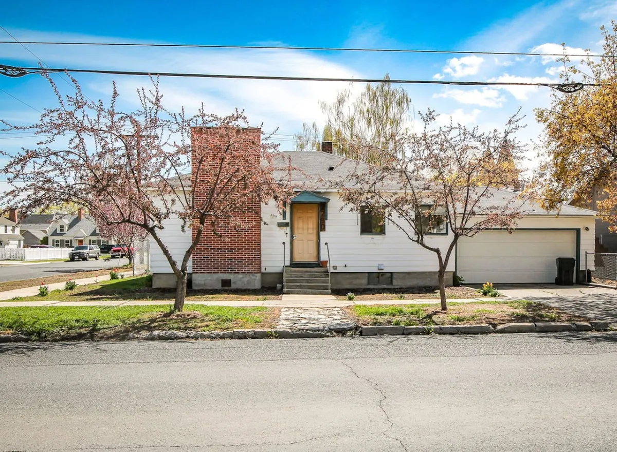 A welcoming exterior view of a home with cherry blossom trees in bloom, a brick chimney, and a stone pathway leading to a charming front door. The setting suggests a peaceful, walkable neighborhood ideal for outdoor strolls.