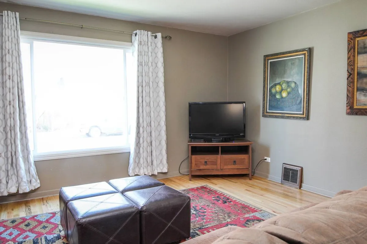 A cozy living room featuring a large window that fills the space with natural light, a sleek television, stylish artwork on the wall, and a coffee table set up for relaxing or entertaining guests. The setting conveys comfort and a welcoming atmosphere for families and friends.